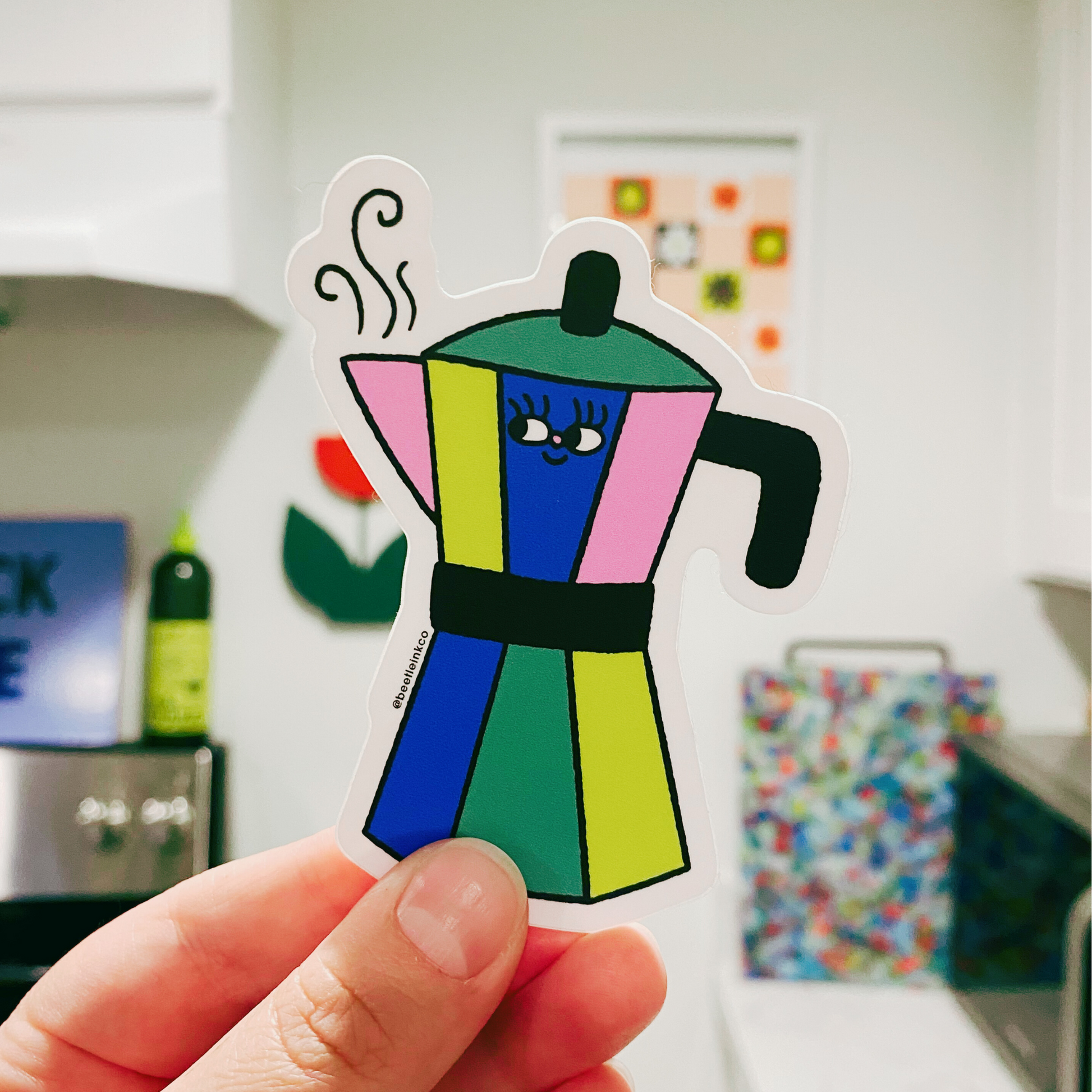 The artist holds up a hand drawn vinyl sticker in the shape of a stovetop espresso maker, with a cartoon smiley face, in her kitchen. 